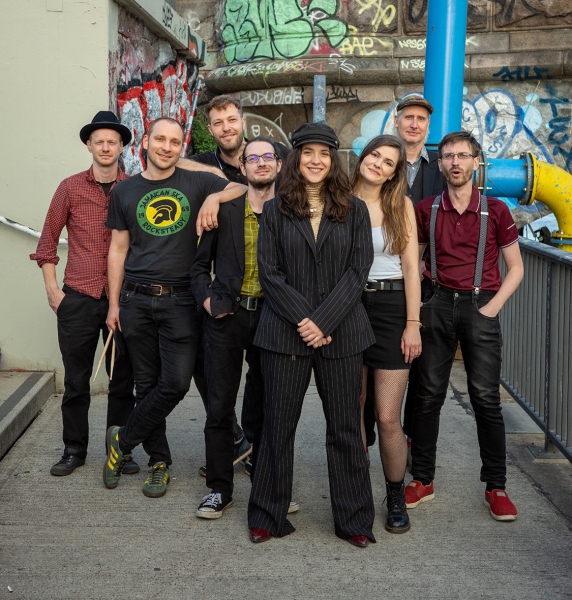 Foto der Band Arte Bella & The Fellas. Eine Gruppe von acht Personen steht auf einem urbanen Gehweg mit Graffiti-Wänden im Hintergrund. Alle Personen schauen freundlich in die Kamera. Die Gruppe ist gemischt, mit Männern und Frauen, alle in lockerer, indi